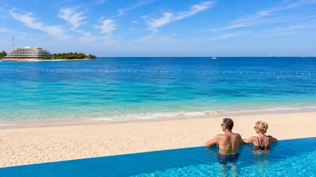 A couple relaxes in an infinity pool overlooking calm turquoise water along the Nassau coastline.