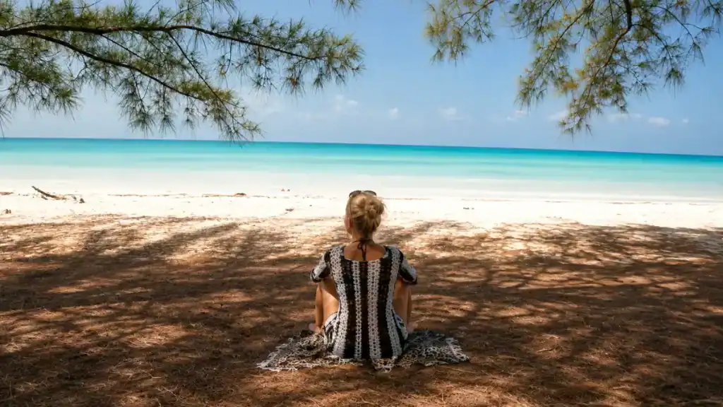 Woman sits under casuarina shade facing the shoreline in Eleuthera