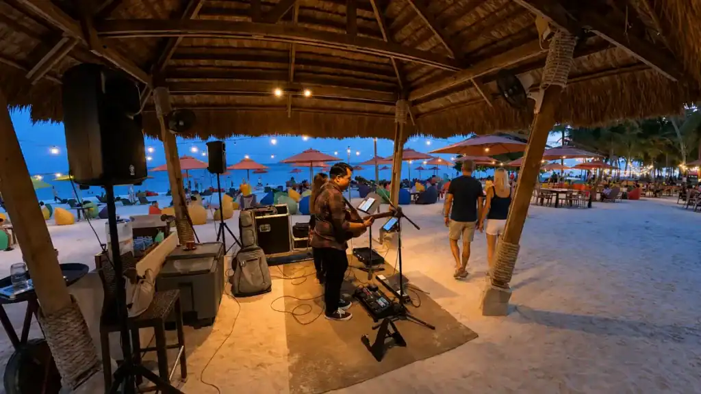 A live guitarist performs under a thatched roof beside beanbags and umbrella lights at dusk on Alona Beach.