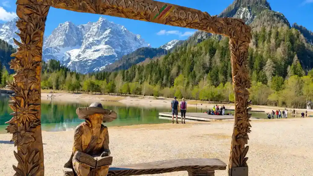 A carved wooden gateway frames snow-capped peaks and a lakeside statue reading a book by the shore.