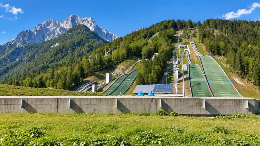 Green synthetic ski jump towers rise against jagged peaks on a summer hillside in Planica.