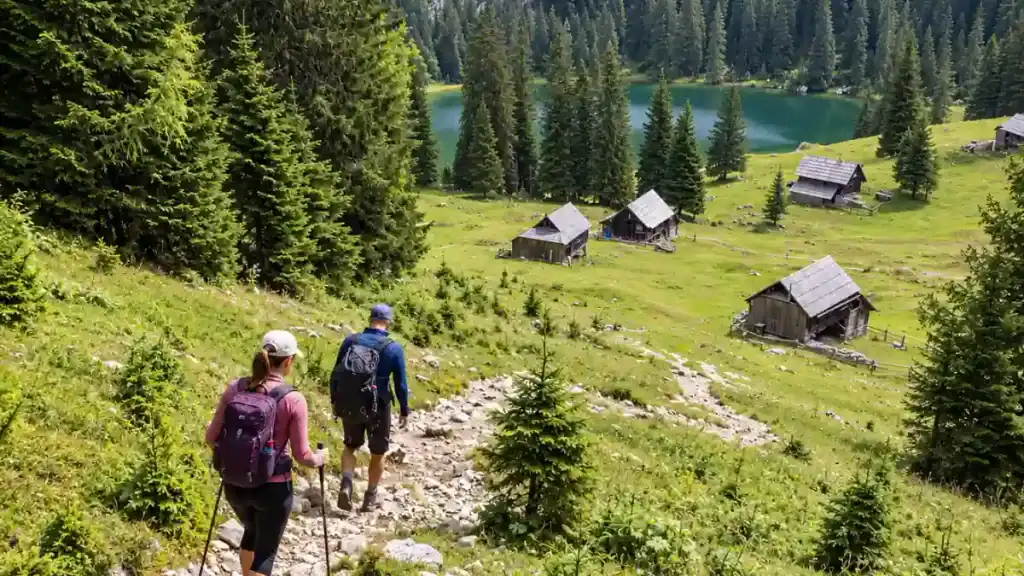 Hikers descend a rocky path toward wooden herders' huts beside an emerald mountain lake.