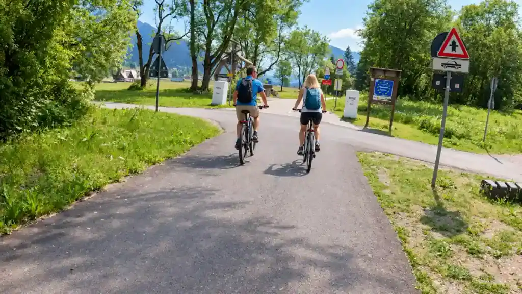 Cyclists follow a paved path past a border marker near Jasna, framed by green hillsides.