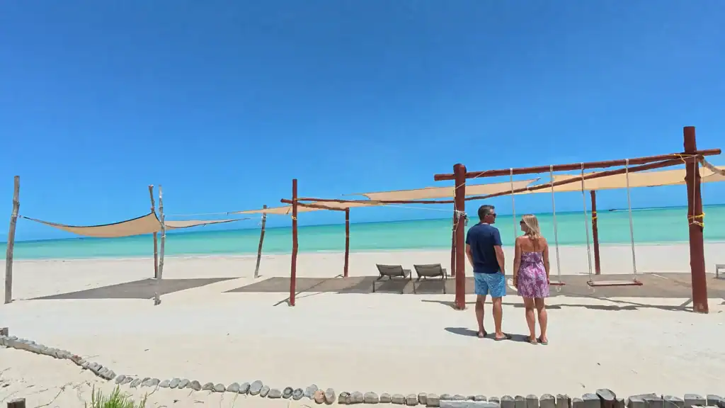 Couple standing beneath wooden beach cabanas questioning is Holbox expensive along turquoise shoreline.