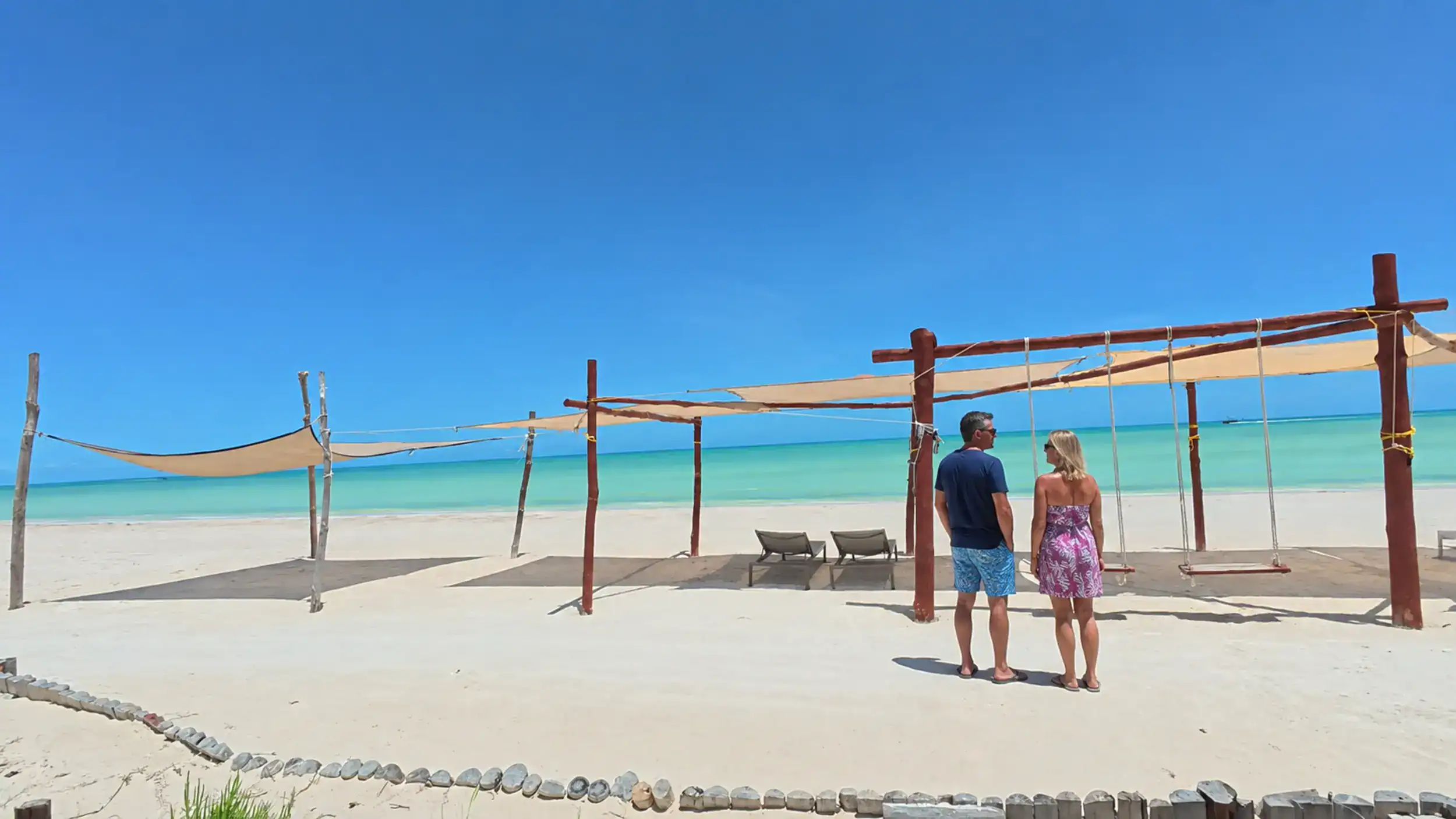Couple standing beneath wooden beach cabanas questioning is Holbox expensive along turquoise shoreline.