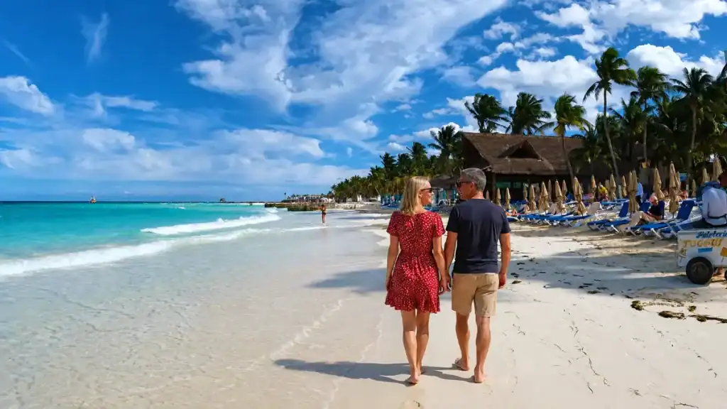 A couple strolls the shoreline near palapa roofs and loungers at Mayan Beach Club.