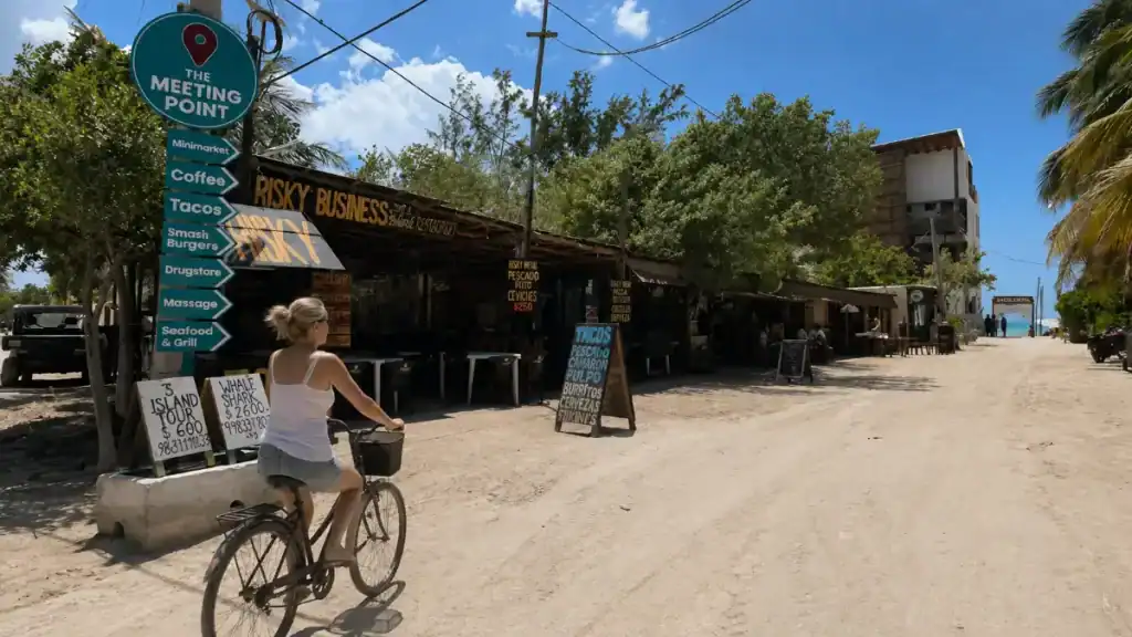 Woman bikes past The Meeting Point sign and Risky Business restaurant on a sandy town road in Holbox.