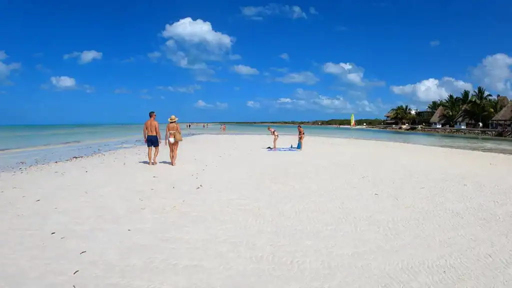 Beachgoers spread across a wide sandbar with palapa roofs visible along the far shore.