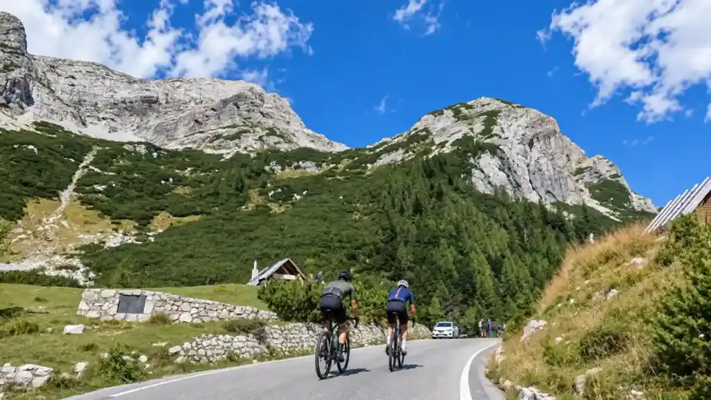 Road cyclists climb toward a memorial stone below the bare limestone ridges near the pass summit