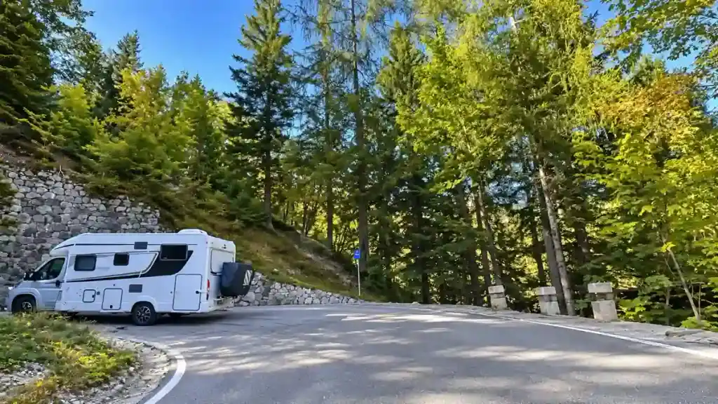 A campervan navigates a stone-walled hairpin bend through forest in the Julian Alps