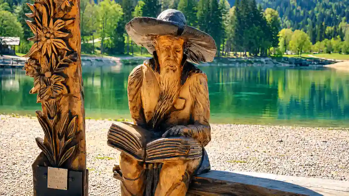 A carved wooden storyteller figure sits beside Lake Jasna holding an open book