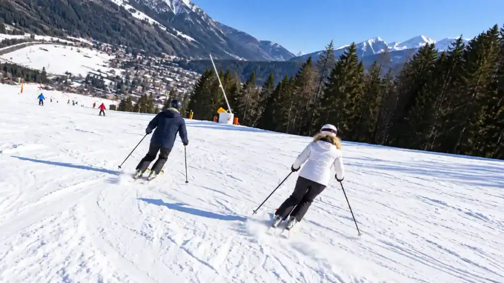 Two skiers lean into parallel turns above a valley town ringed by snow-capped peaks.