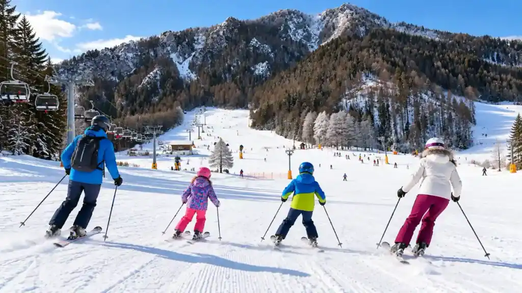 A family of four pauses mid-run, poles planted, above a tree-lined chairlift corridor in the Julian Alps.