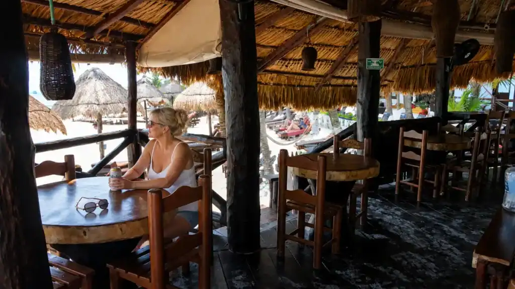 Woman sits with a drink at a wooden table inside a thatched-roof Holbox beach bar.