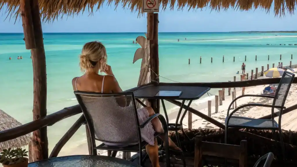 Woman sits at a beachfront table under a palapa, overlooking swimmers in shallow turquoise water.