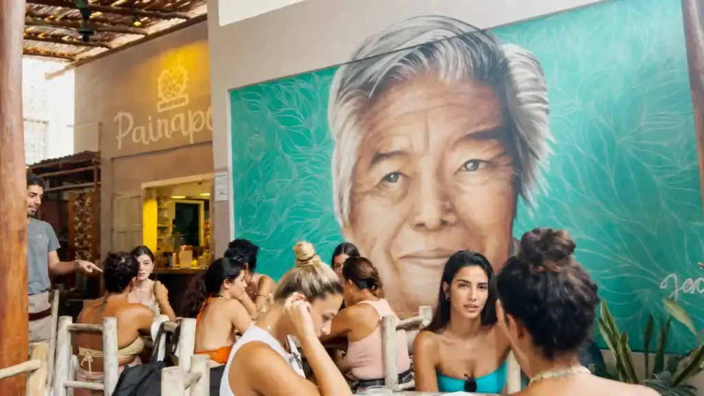 Friends gather at a table inside Painapol café beneath a large mural of a man's portrait.