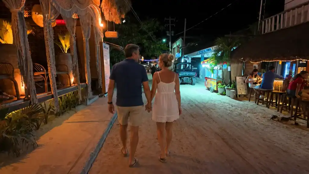 Couple strolls hand in hand down a lantern-lit sandy street past bars and a golf cart at night.